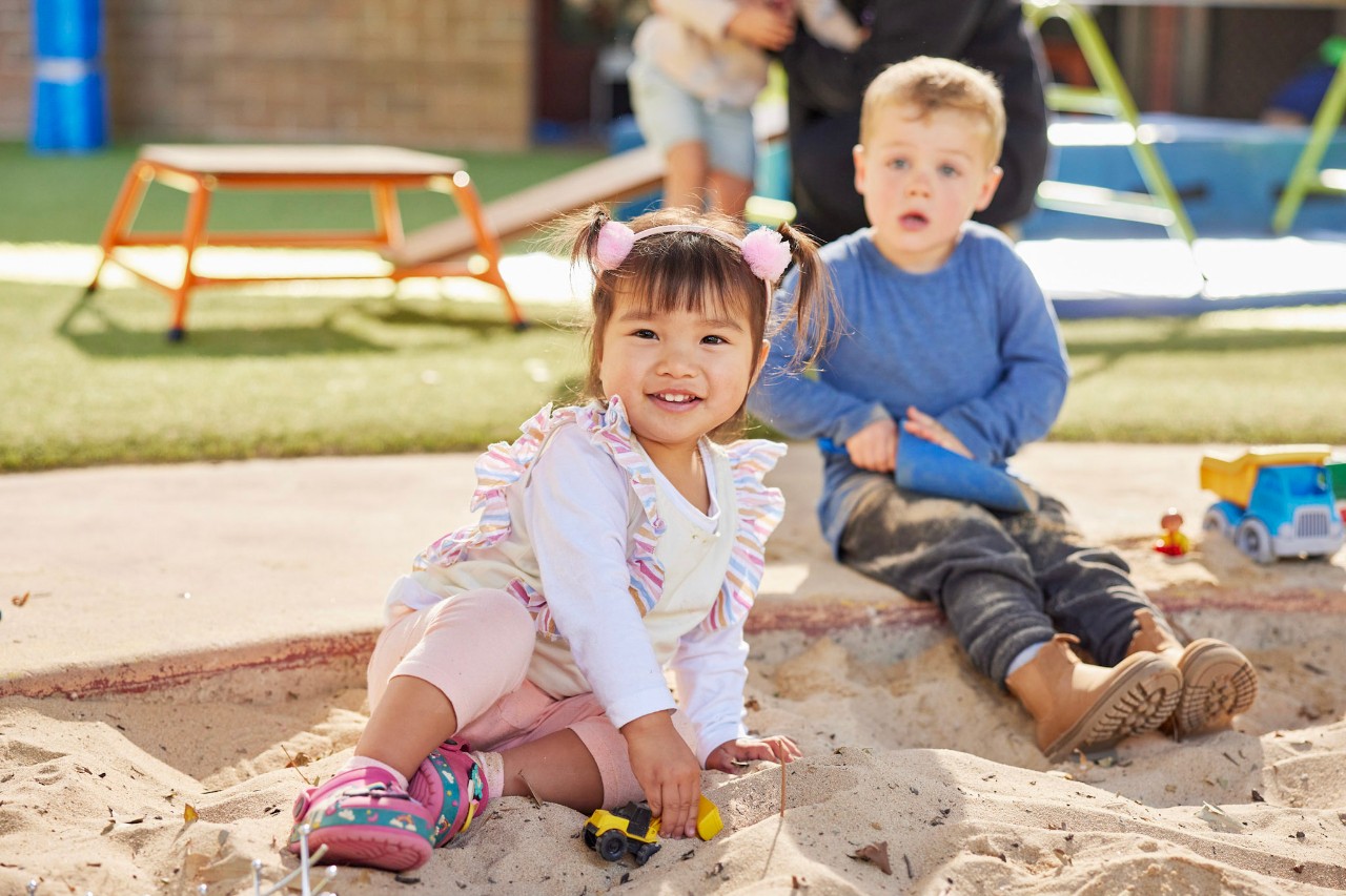 Two children sit in a sand pit holding toys.