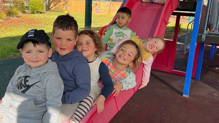 Photo showing six children sitting one behind the other on the bottom part of a slide in a playground.