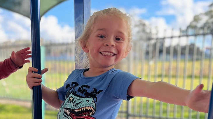 Photo of young girl with blonde hair from the waist up smiling wearing a blue t-shirt reaching from one pole to the next on play equipment.