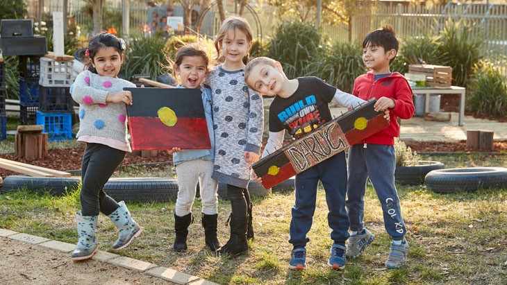 three girls and two boys stand outside in an ECEC service together smiling. The two girls furthest on the left are holding a hand painted Aboriginal Flag. The boy second from the right is holding a rectangular sign hand painted with the word Darug in the middle done in traditional dot painting style with two Aboriginal flags painted on either side of the word.