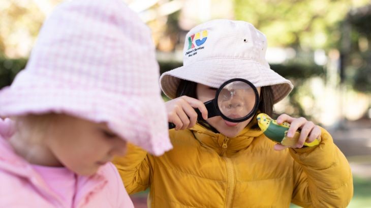 Two children outside at an early childhood education and care service. One child looks at a toy bird through a magnifying glass.