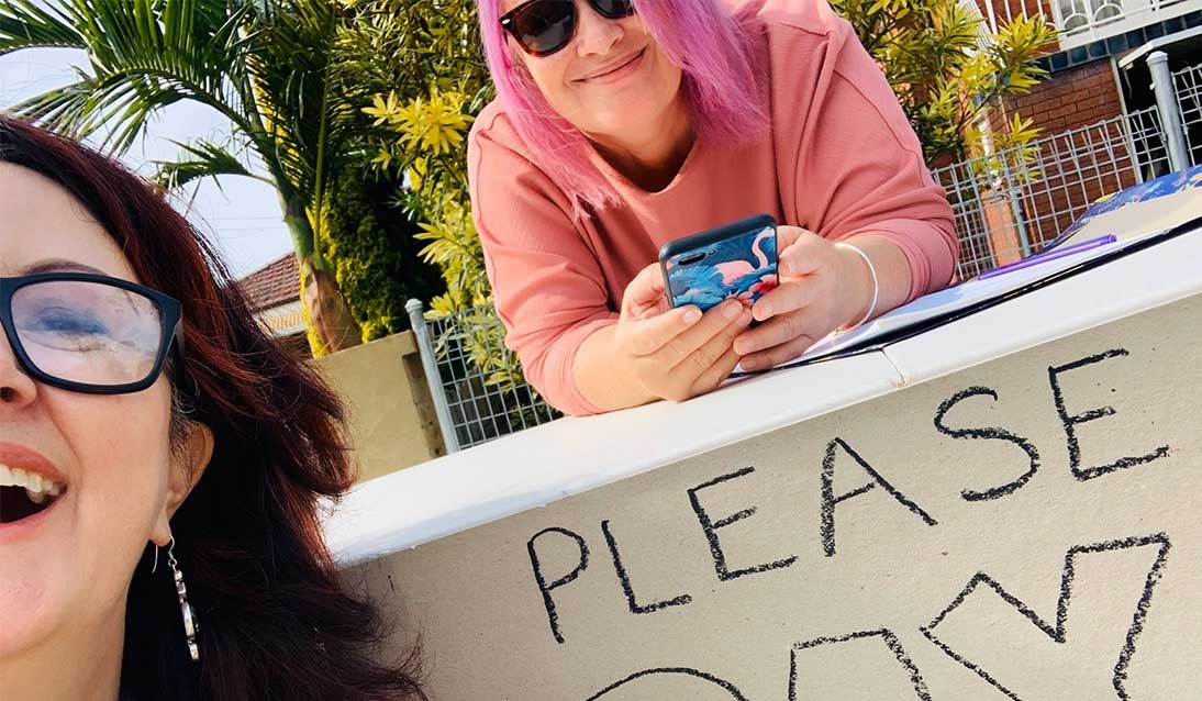 Two women smiling at a table with a sign reading please pay.