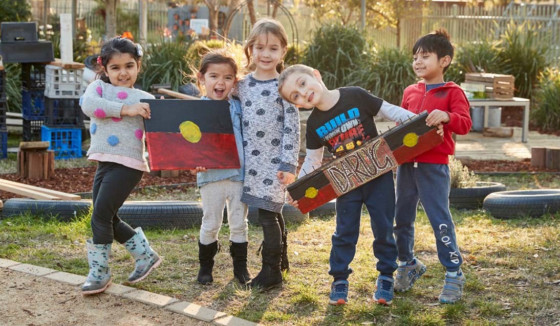 Preschool students stand outside holding Aboriginal Flags