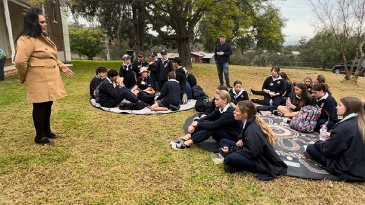 School students listen to an Aboriginal Elder talk about the history of the Cootamundra Girls Home while sitting eating lunch.