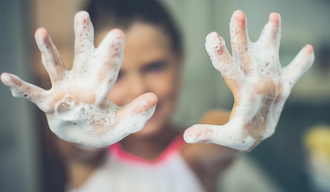A young girl holds up soap covered hands.