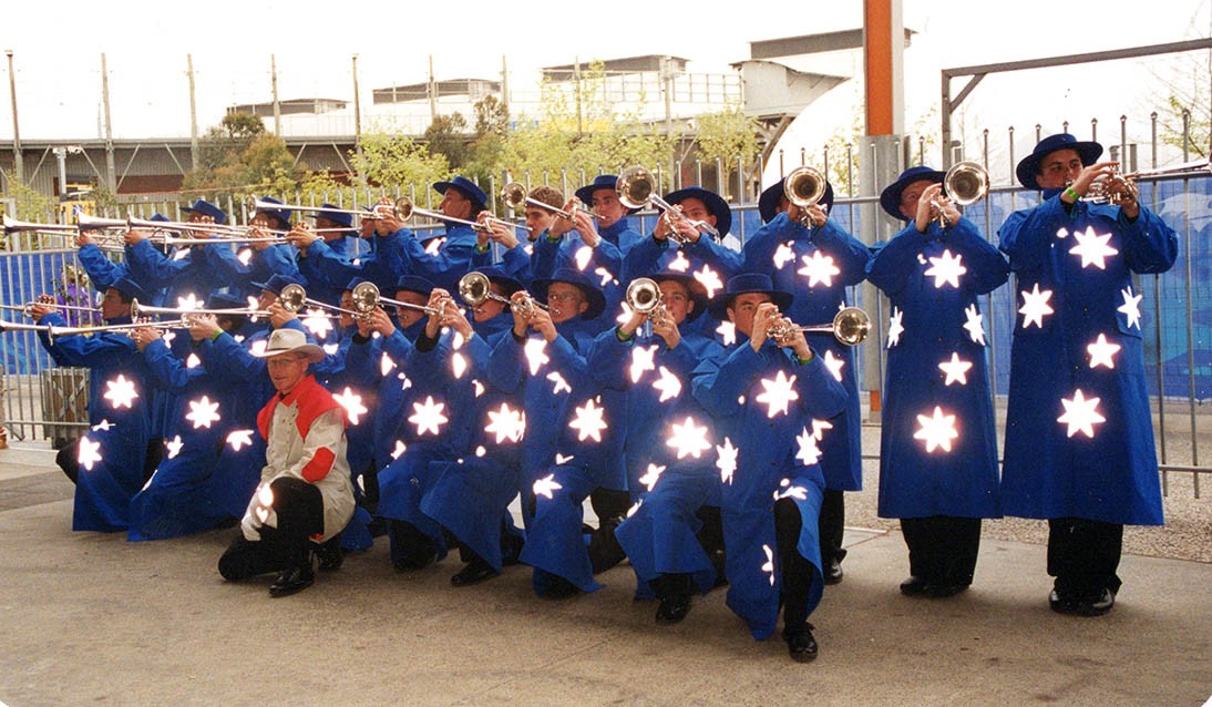 Group of people holding trumpets and wearing blue outfits. Man in red outfit kneels in front of group.