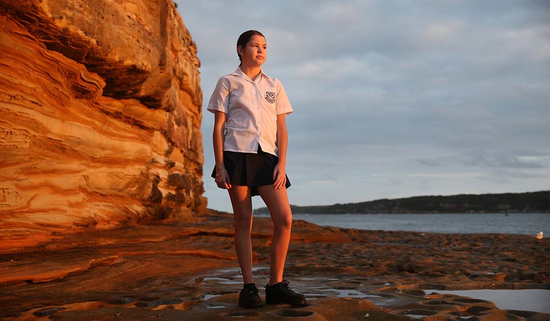 A teenage girl stands on a rock shelf overlooking the water.