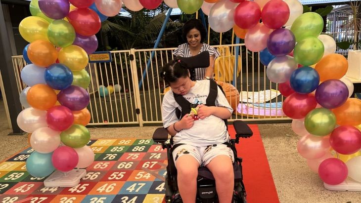 A student in a wheelchair with a teacher on a red carpet under a balloon arch.