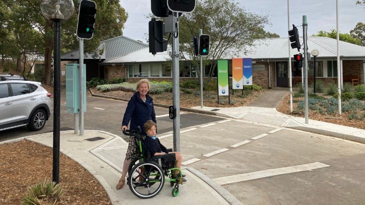 A woman and a boy in a wheelchair standing on a footpath at a set of traffic lights.