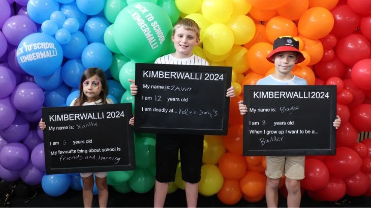 Three children standing in front of coloured balloons holding placards.