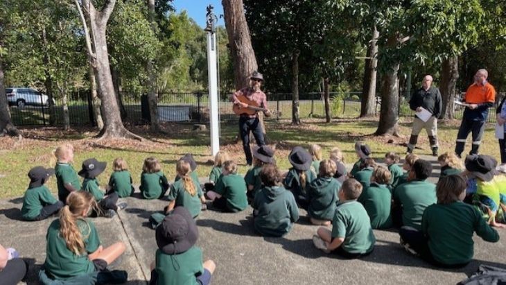 Students sitting on the ground watching a man play guitar.