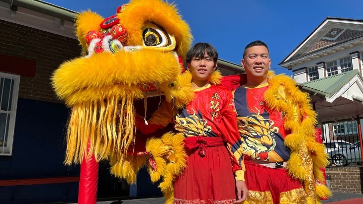 Two people in costume in front of a Chinese lion.