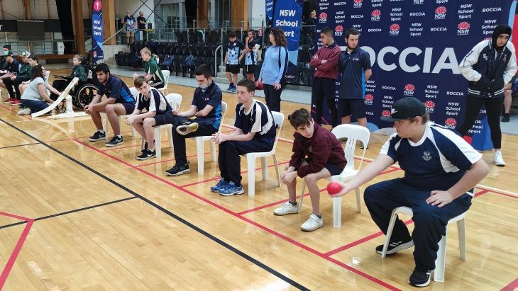 Students seated in chairs on a wooden court holding balls.