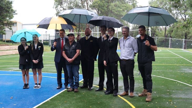 Students and adults holding umbrellas standing on an artificial grass court.