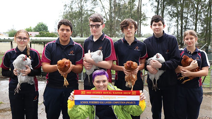 Six people holding chickens stand in a line with a woman holding a ribbon crouched in front of them