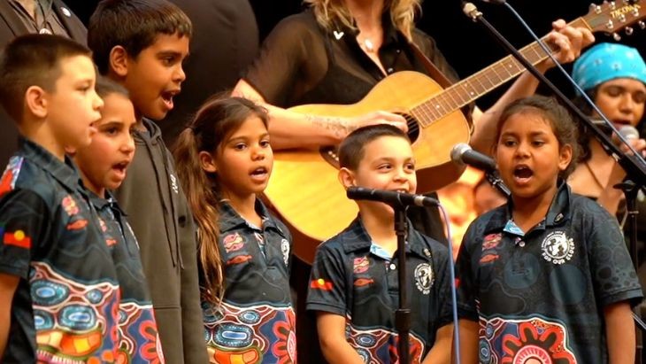 Students dressed in colourful T-shirts with Aboriginal designs singing into microphones.