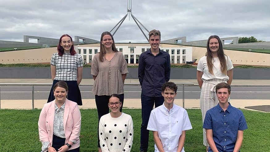 A group of eight people in two rows with Parliament House in the background.