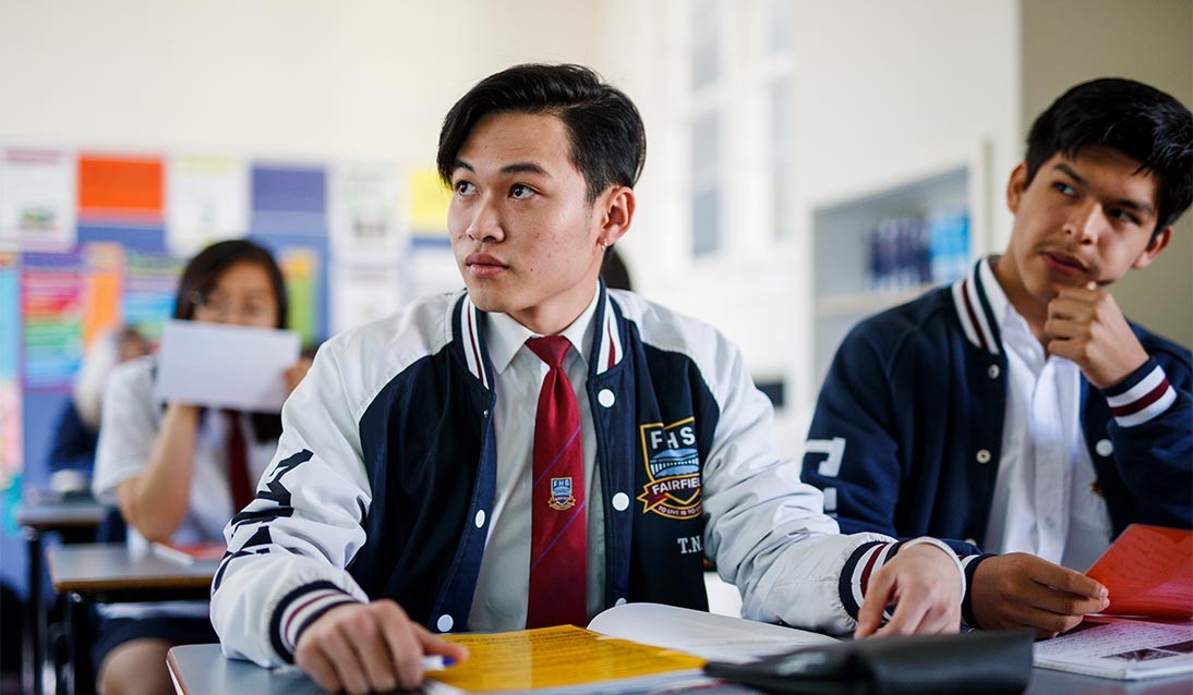 Two male students wearing school uniform sit in class at a desk, looking to the side of the camera.
