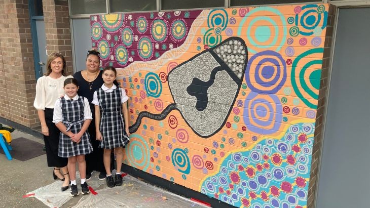 Two women and two students standing in front of a wall with a mural painted on it.