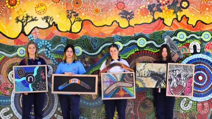 Students holding paintings in front of a wall mural.