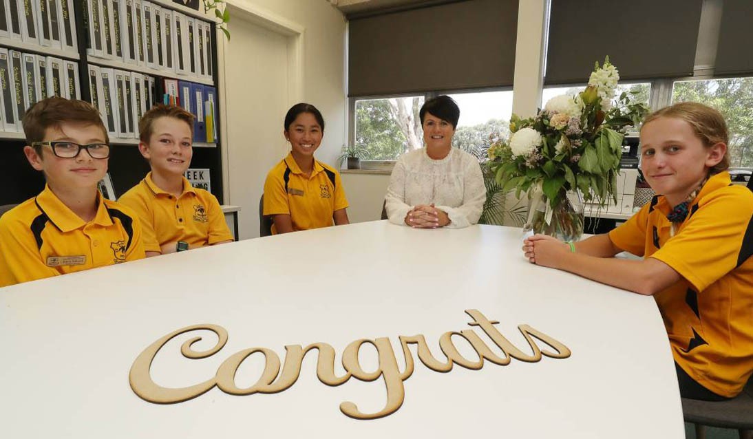 Four students wearing yellow school uniform and adult wearing white sit around a white table indoors with the text Congrats displayed on the table.