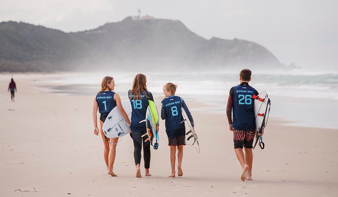 Four students walking away from the camera on the beach with surfboards