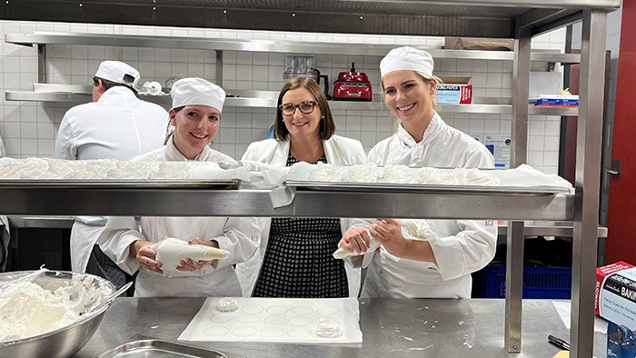 Two girls dressed in chefs uniform stand either side of the Education Minister in a kitchen