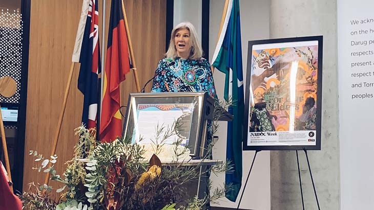 A woman speaking at a lectern standing next to a painting and in front of some flags.