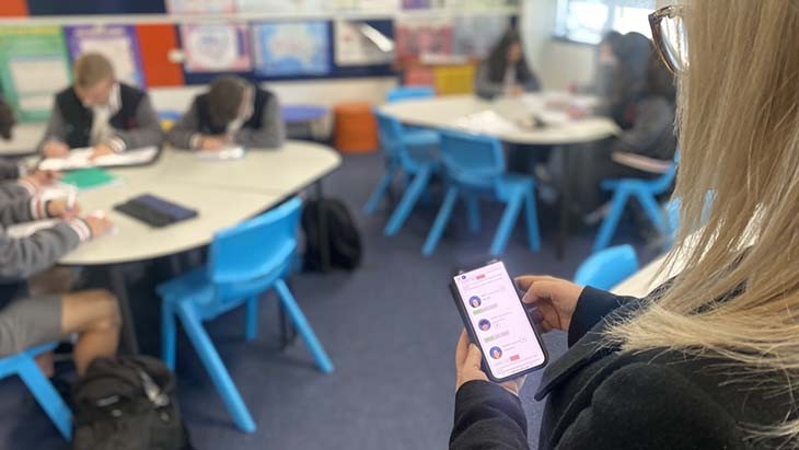 A teacher looks at an image on a mobile phone while students work in the background.