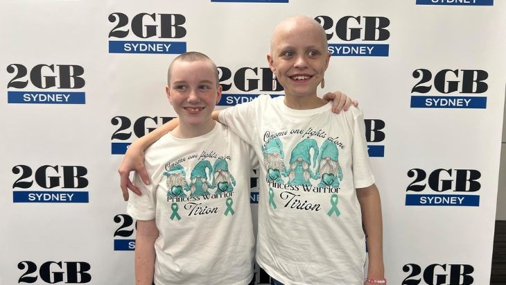 Two girls with shaved heads standing in front of a media banner.