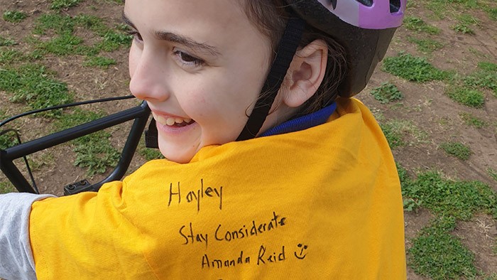 A young girl on a bicycle shows off an autograph on the back of her yellow shirt.