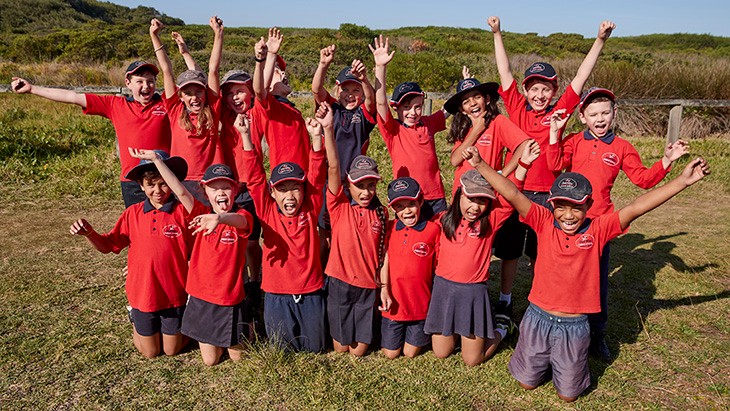 A large group of primary students with their hands in the air near a beach.