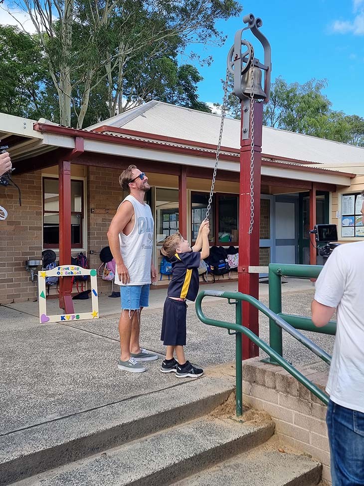 A little boy ringing a school bell while his dad watches.