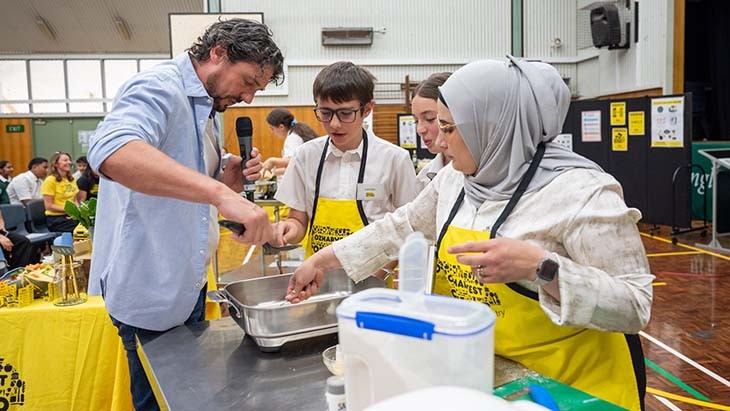 A man dips a spoon into a mixing bowl while students look on.