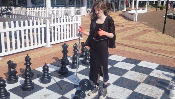 A girl in costume holding a sword and standing on a giant chess board.