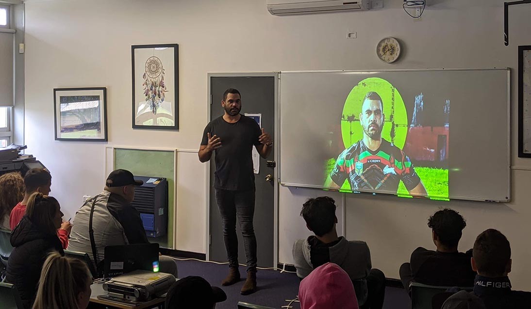 Man stands in front of classroom with image of footballer projected onto whiteboard. Students sit on chairs in classroom.