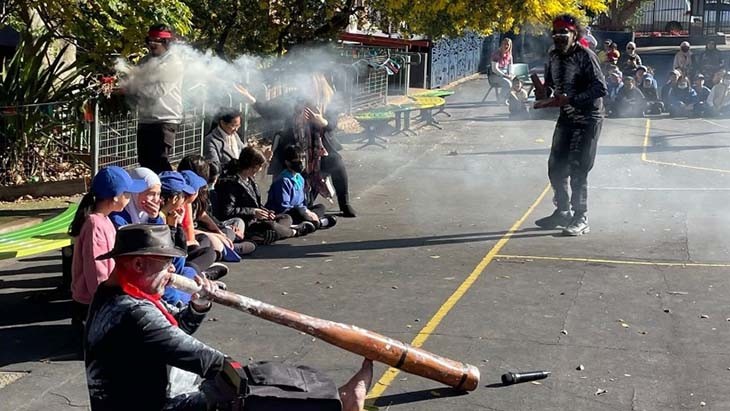 A man plays a didgeridoo while another man performs a smoking ceremony.