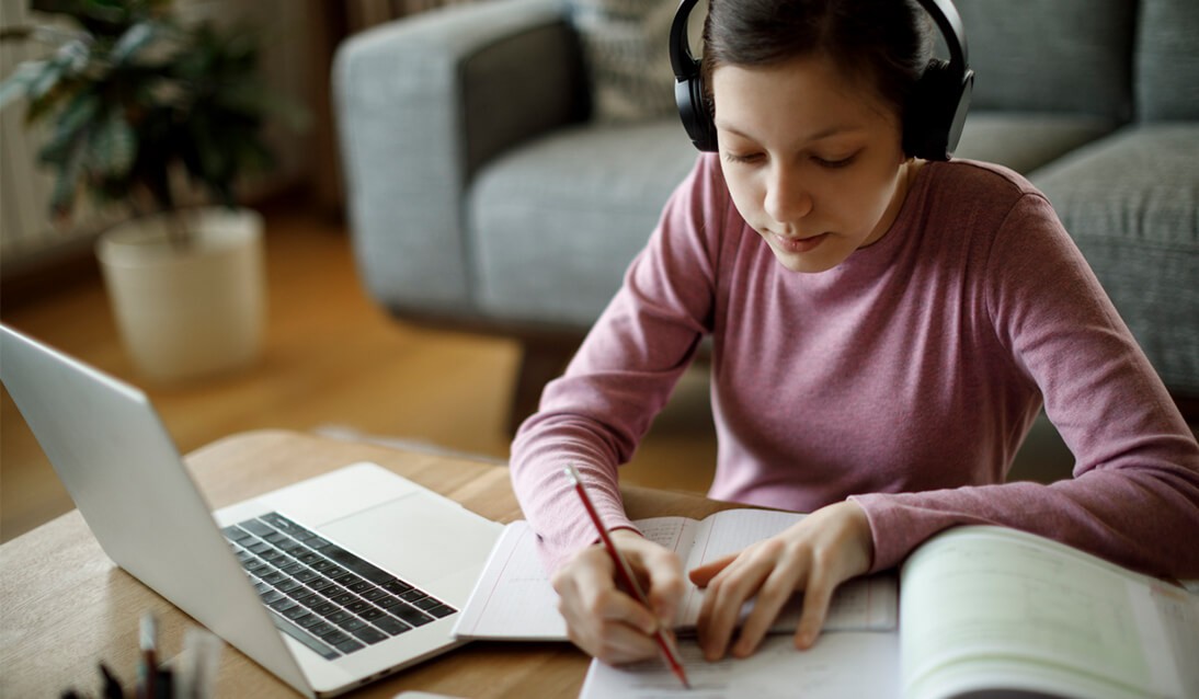 Student wearing headphones sitting at desk at home working on computer and paper book