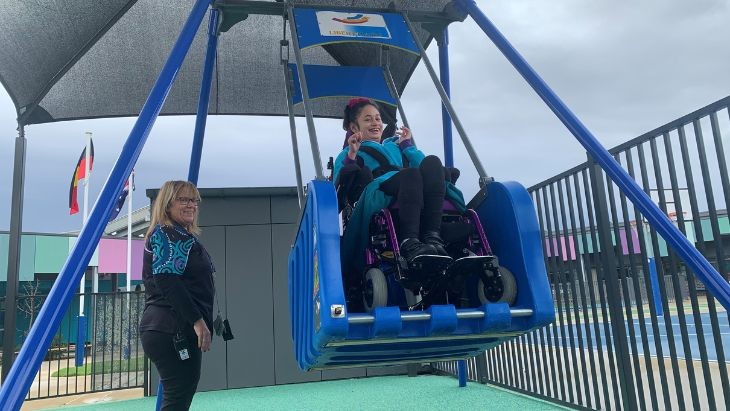 A girl in a wheelchair on a swing with a lady standing beside her.