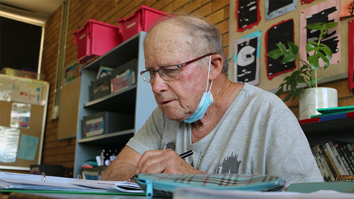An elderly man is taking notes, sitting at a desk in a classroom.