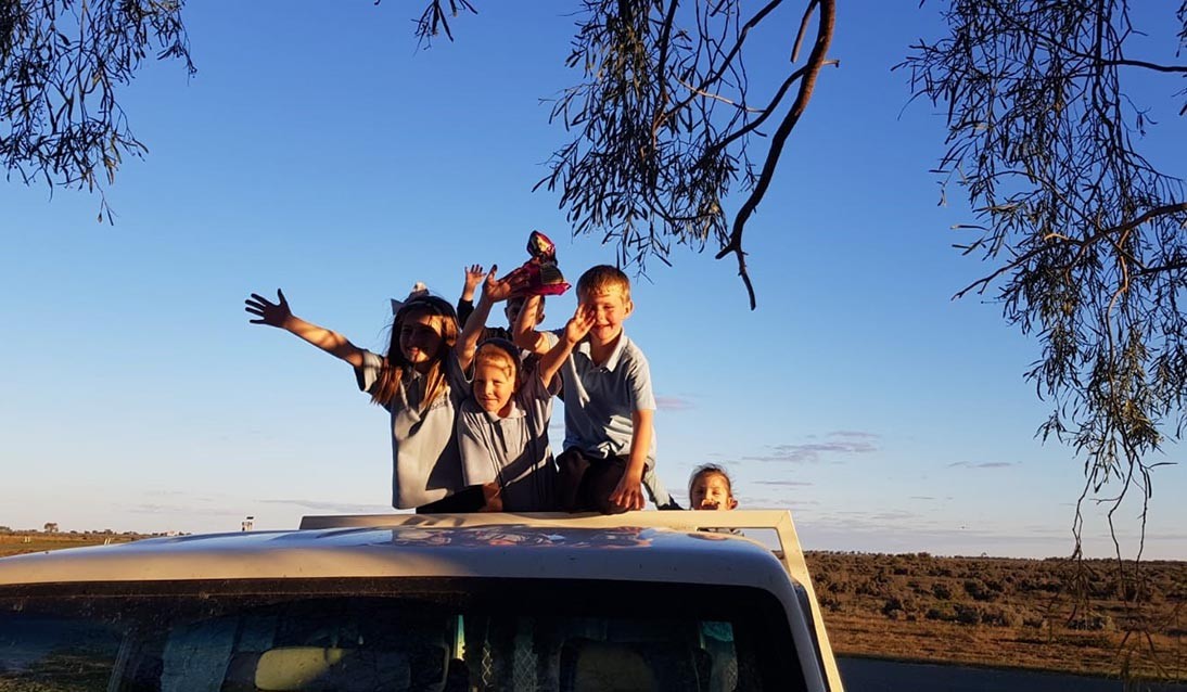 Small group of students strand on back of car in middle of outback. Blue sky is in the background with trees hanging down on side of photo.