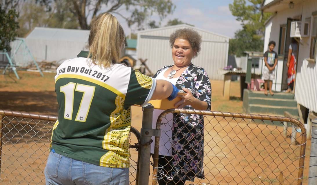 A woman wearing a Menindee Central School shirt hands a box of food over a fence to another lady.
