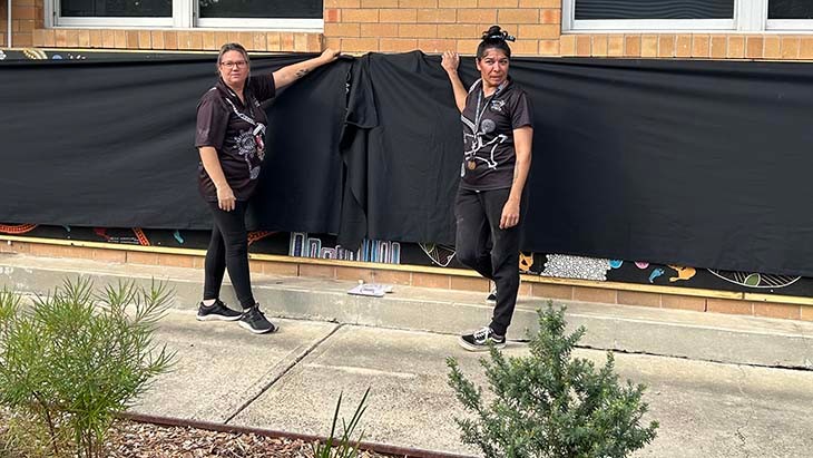 Two women hold the corners of a black banner on a wall.