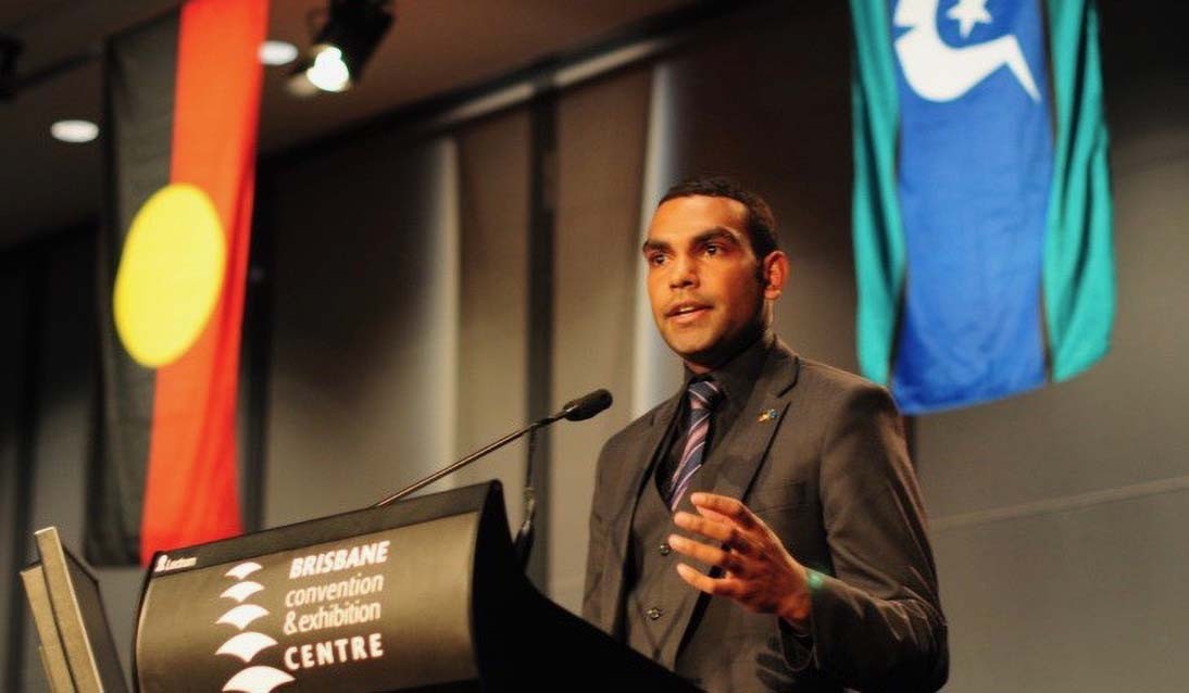 Man wearing a suit stands in front of a lecturn and microphone. Aboriginal flag and Torres Strait Island flag hangs from the ceiling behind him.