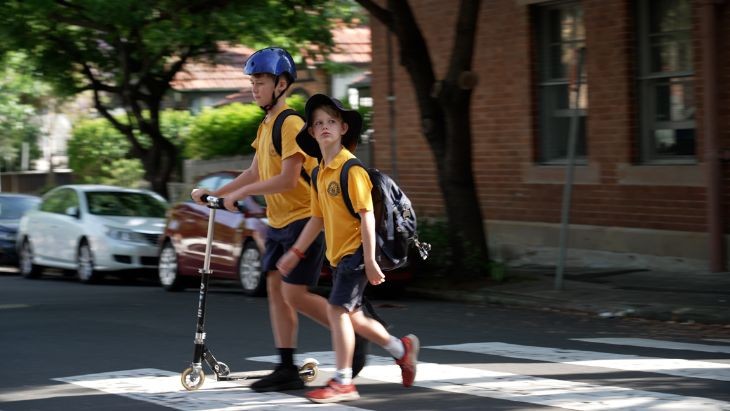 Two students crossing the road. One is pushing a scooter.