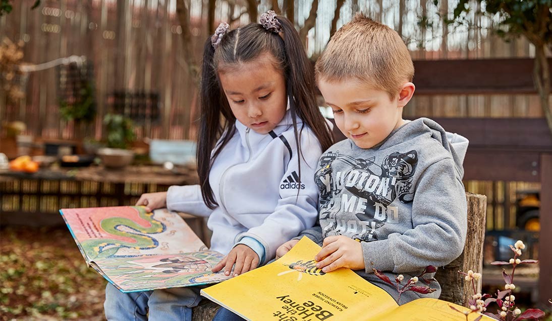 Two children sitting in a playground reading picture books.