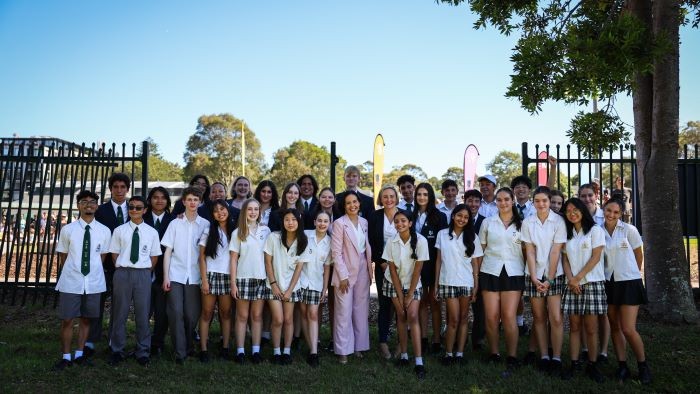 A group of students surrounding an adult in front of a gap in a fence