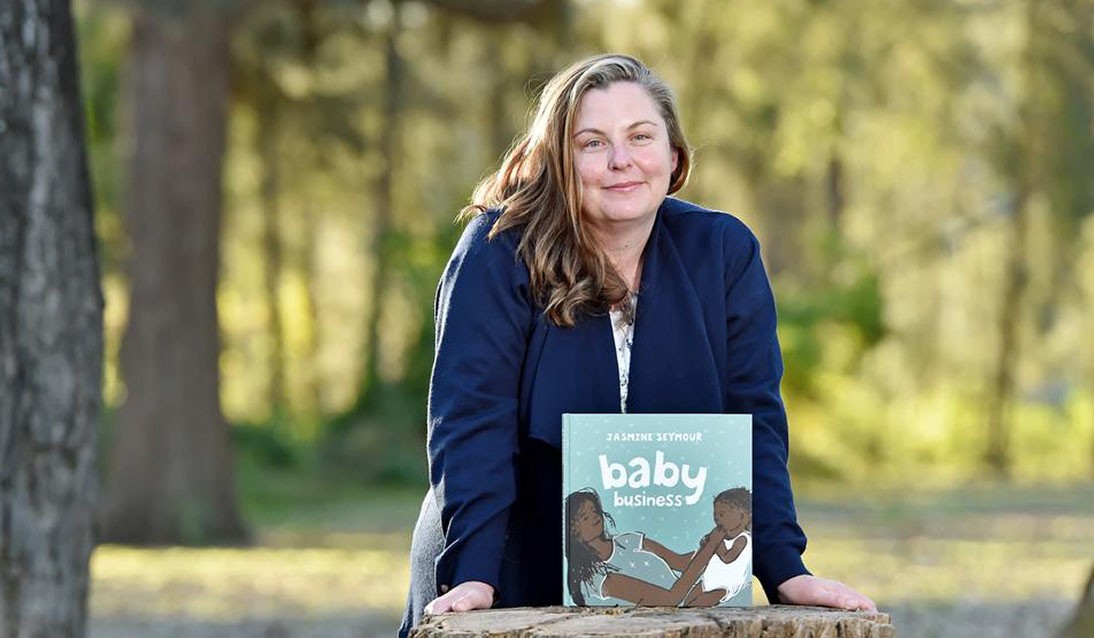 Woman wearing navy blue jacket leans against a tree stump outside with picture book Baby Business resting on top of the tree stump. Trees are in the background.