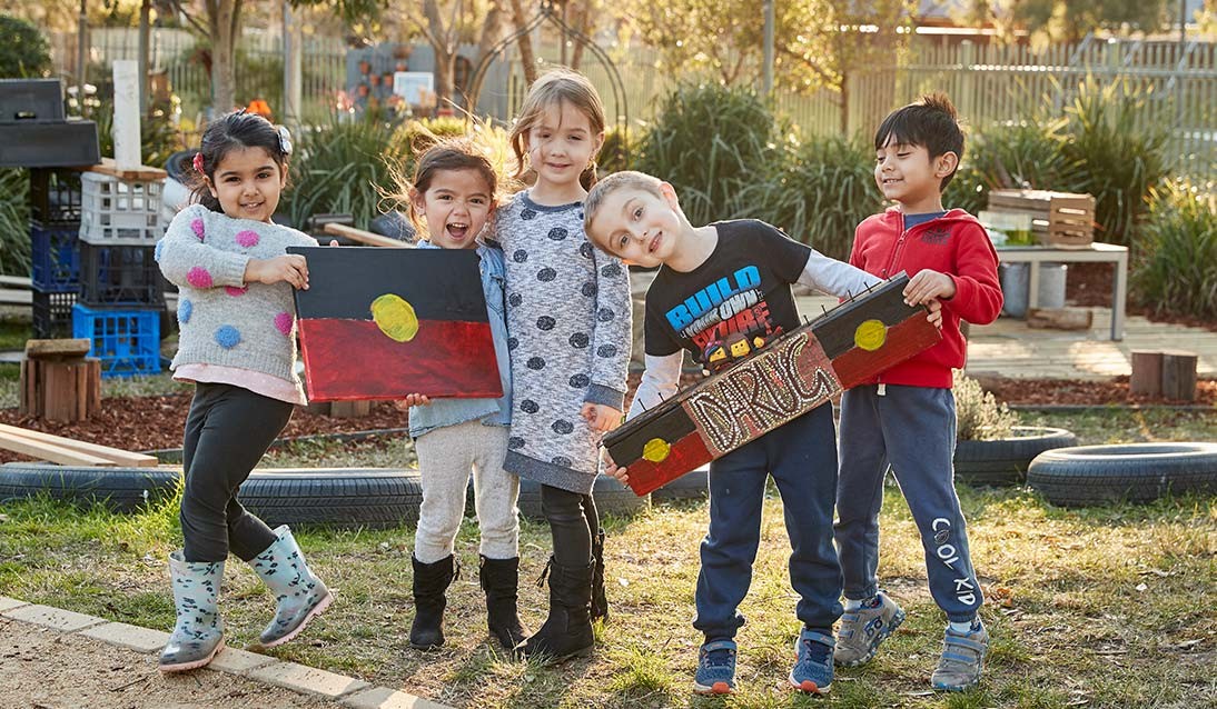 A group of preschool students holding painted Aboriginal flags and a sign with the word Darug.