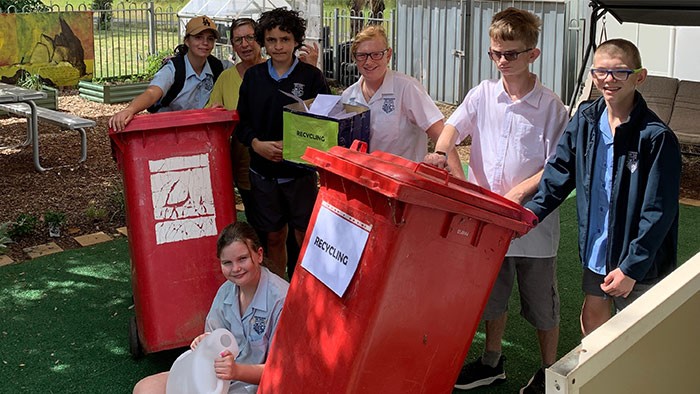 A group of students and one teacher standing near red recycling bins.